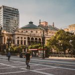 Pedestres caminham por uma praça diante de edifícios modernos e do Theatro Municipal de São Paulo.