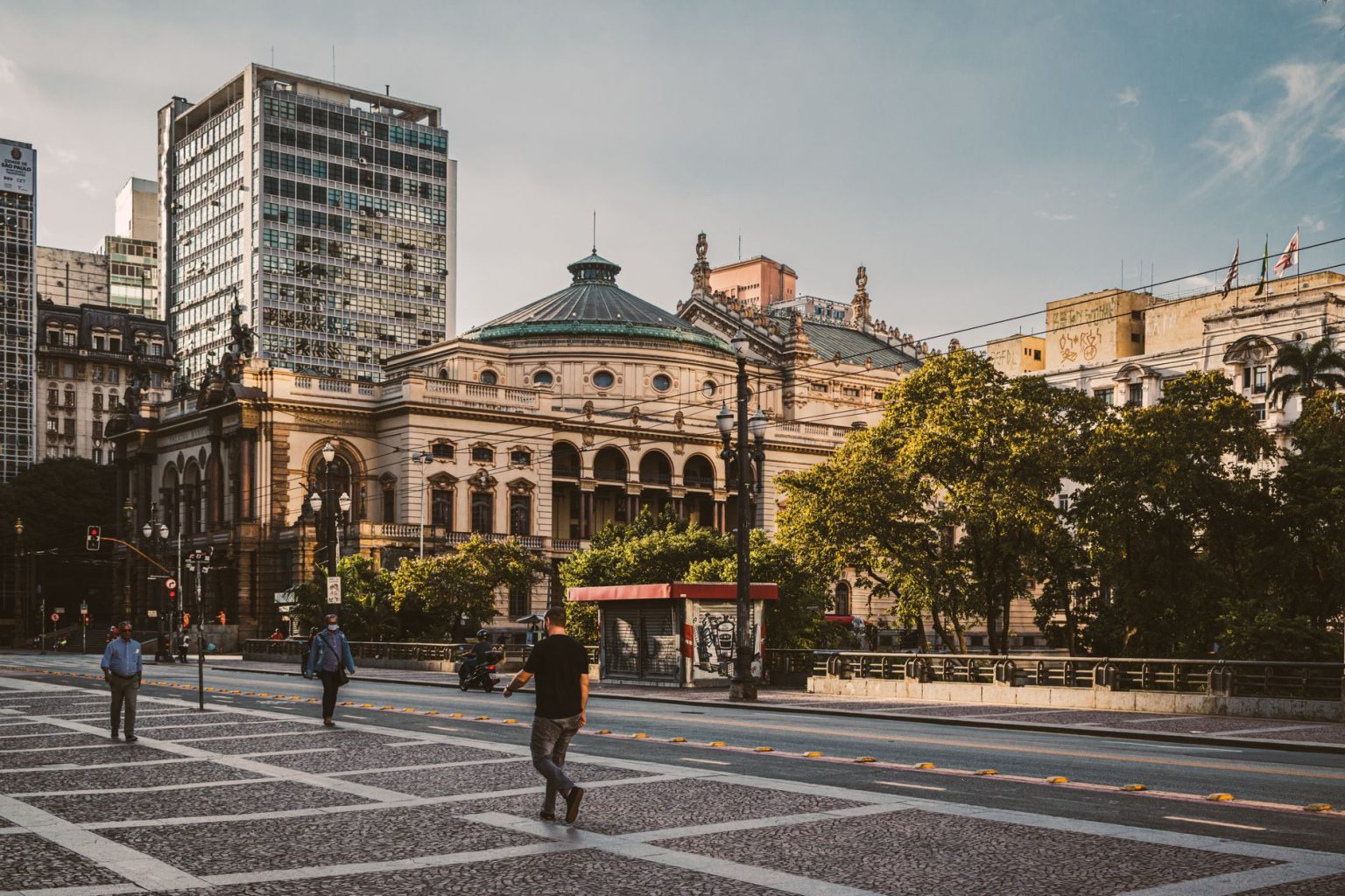 Pedestres caminham por uma praça diante de edifícios modernos e do Theatro Municipal de São Paulo.