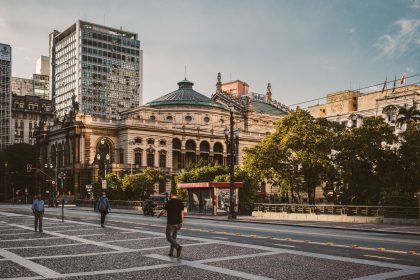 Pedestres caminham por uma praça diante de edifícios modernos e do Theatro Municipal de São Paulo.