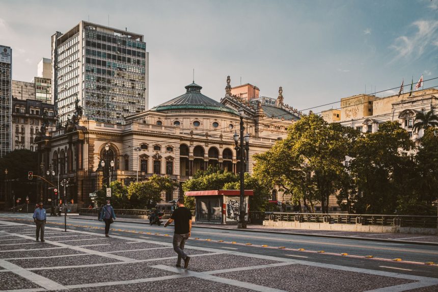 Pedestres caminham por uma praça diante de edifícios modernos e do Theatro Municipal de São Paulo.